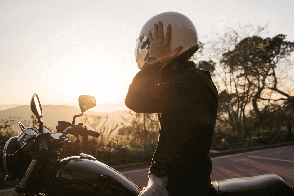 A motorcyclist taking off their white helmet while standing beside their motorcycle during sunset.
