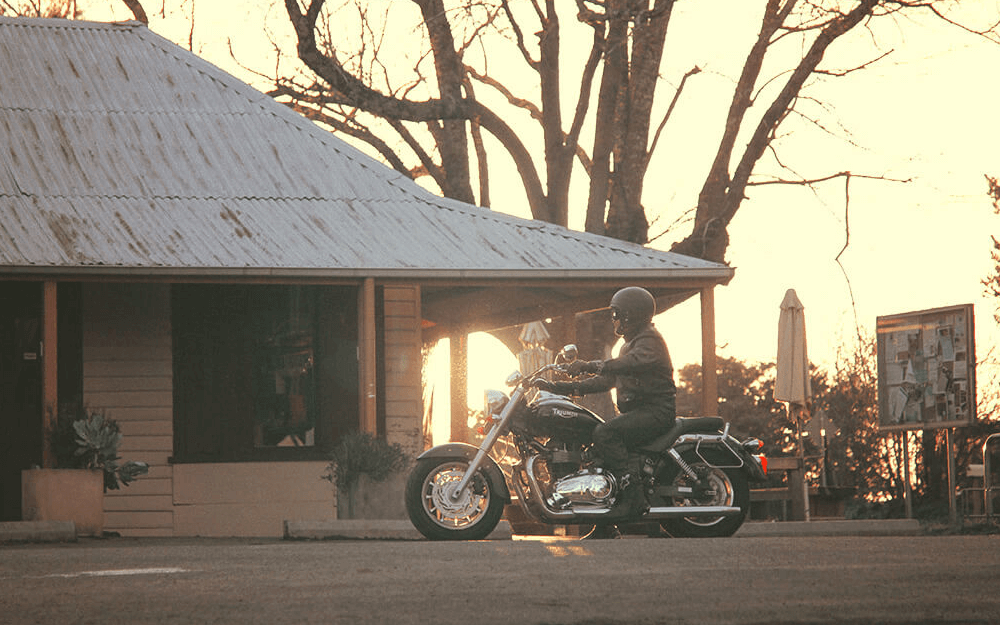 A motorcyclist riding past a rustic wooden building at dusk.